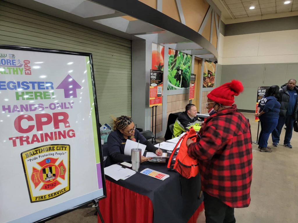 A Baltimore City Fire Department table at a community health expo offering hands-only CPR training registration. A large sign with the department logo stands in the foreground, while staff assist attendees at the booth. A woman in a red plaid jacket and hat is speaking with a representative.
